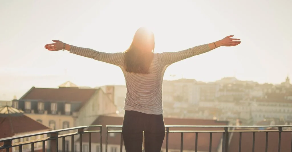 A woman standing with here arms wide open outside, catching some sunshine.